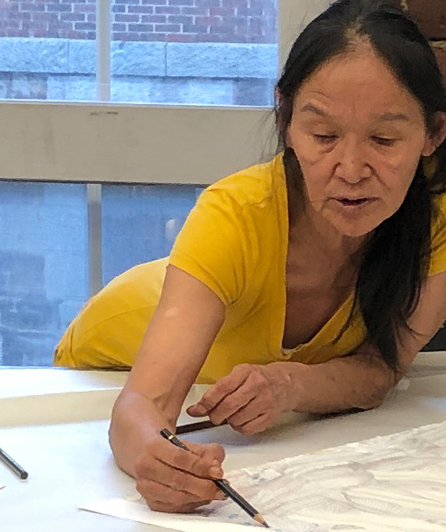 Colour photograph of artist Shuvinai Ashoona in a yellow t-shirt bent over a table drawing on paper in brown pencil crayon. A window is behind her where you see a neighbouring brick building. 