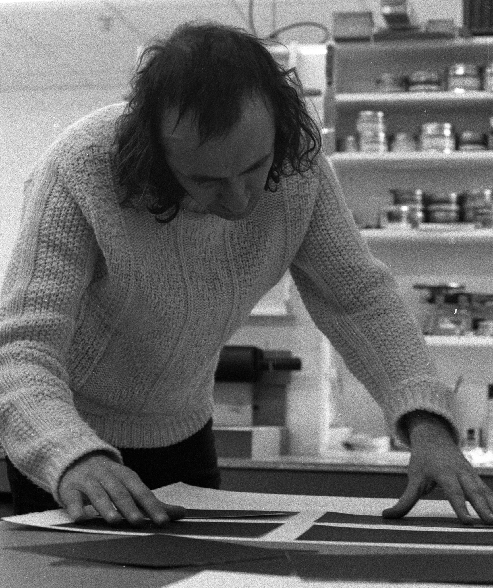 Black and white photo of artist Vito Acconci, with shoulder length hair wearing a cable knit sweater adjusting prints in the print studio. A shelf of inks and chemicals is behind him.